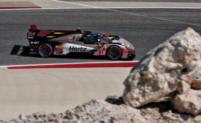 Prototype race car (Cadillac DPi) with Hertz sponsor speeding around a turn on an asphalt track, rock foreground on the right side.
