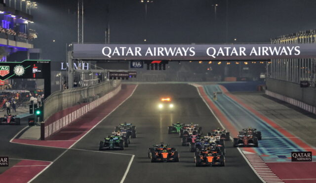 Nighttime Formula 1 race start: multiple cars line up on a illuminated track beneath a large Qatar Airways banner.",