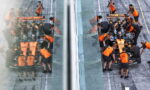Pit crew in orange uniforms service a Formula 1 car at the pit stop, viewed through glass walls with the car split by a transparent divider.