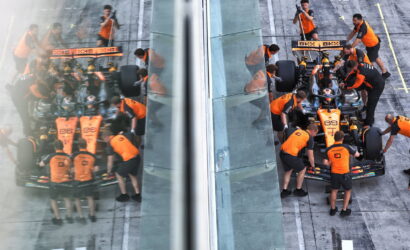 Pit crew in orange uniforms service a Formula 1 car at the pit stop, viewed through glass walls with the car split by a transparent divider.