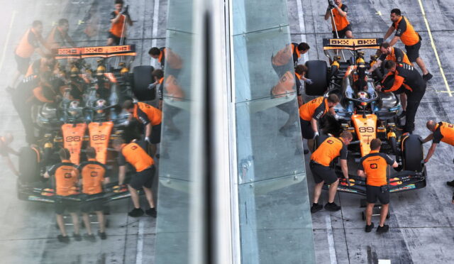 Pit crew in orange uniforms service a Formula 1 car at the pit stop, viewed through glass walls with the car split by a transparent divider.