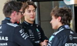 Three Mercedes Formula 1 team members in black racing suits talk in a pit area, one appears to shake hands with another while a third watches.