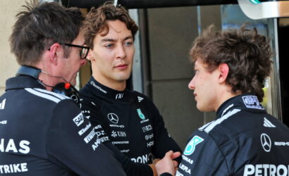 Three Mercedes Formula 1 team members in black racing suits talk in a pit area, one appears to shake hands with another while a third watches.