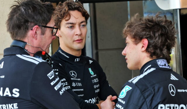 Three Mercedes Formula 1 team members in black racing suits talk in a pit area, one appears to shake hands with another while a third watches.