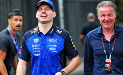 Smiling race car driver in a blue Red Bull Racing shirt and cap walks beside a man in a dark polo with a lanyard badge.