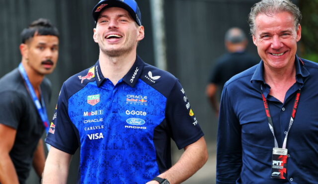 Smiling race car driver in a blue Red Bull Racing shirt and cap walks beside a man in a dark polo with a lanyard badge.
