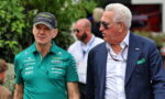 Two men at a public event: one in a green Formula One team polo, cap and badges, the other in a navy blazer and sunglasses.