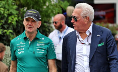 Two men at a public event: one in a green Formula One team polo, cap and badges, the other in a navy blazer and sunglasses.
