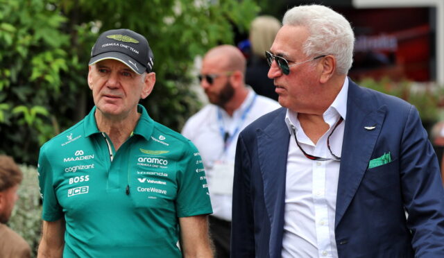 Two men at a public event: one in a green Formula One team polo, cap and badges, the other in a navy blazer and sunglasses.