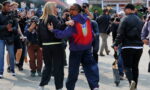 Two people dancing arm-in-arm among a crowd of photographers at an outdoor event.