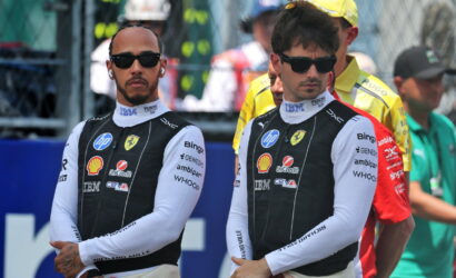 Two Formula 1 drivers in black-and-white Ferrari racing suits wearing sunglasses, standing with arms crossed at a track event, logos visible on suits (Shell, HP).