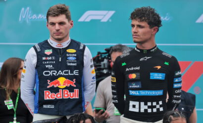 Two Formula 1 drivers stand side by side at a motorsport event, the left in an Oracle Red Bull racing suit and the right in a McLaren suit with sponsor logos, against a Miami F1 backdrop.