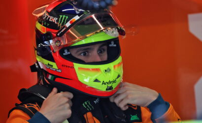 Close-up of a race car driver in an orange suit, adjusting his neon-green helmet with Monster Energy branding.