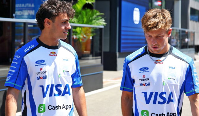 Two young men in blue and white racing jerseys with Visa logos stand on a sunny city street, looking at each other.