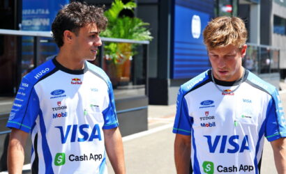 Two young men in blue and white VISA racing jerseys stand on a sunny city street, talking to each other.