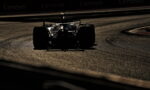 Silhouette of a Formula race car navigating a left-hand curve on a dimly lit race track with the asphalt glow visible in the foreground and background banners faintly seen at the top edge.