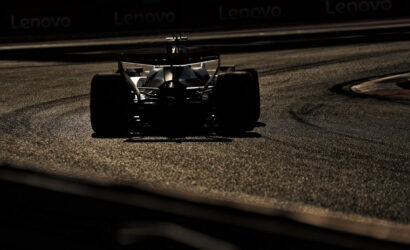 Silhouette of a Formula race car navigating a left-hand curve on a dimly lit race track with the asphalt glow visible in the foreground and background banners faintly seen at the top edge.