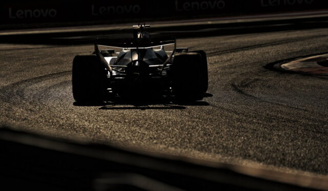 Silhouette of a Formula race car navigating a left-hand curve on a dimly lit race track with the asphalt glow visible in the foreground and background banners faintly seen at the top edge.