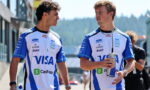 Two men in matching blue-and-white race jerseys walk together outdoors, chatting, each holding a water bottle or canister on a sunny day.
