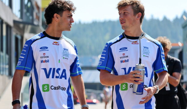 Two men in matching blue-and-white race jerseys walk together outdoors, chatting, each holding a water bottle or canister on a sunny day.