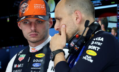 Two Red Bull Racing crew members in a garage, one wearing an orange cap, discussing strategy near equipment.