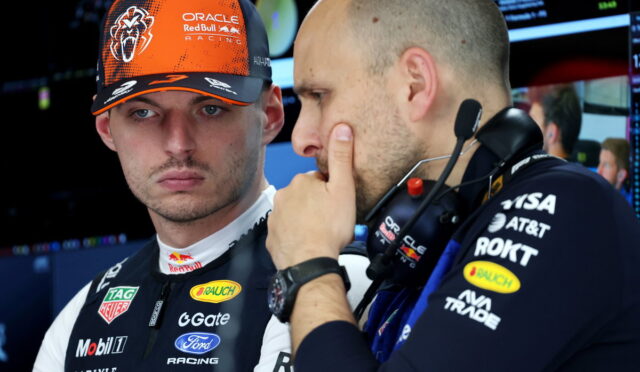 Two Red Bull Racing crew members in a garage, one wearing an orange cap, discussing strategy near equipment.
