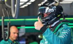 Race car driver in a teal team suit wearing a helmet, giving a thumbs-up in the pit garage.