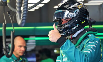 Race car driver in a teal team suit wearing a helmet, giving a thumbs-up in the pit garage.