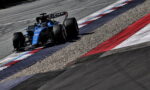 Blue Formula 1 race car rounds a corner on a track, riding the red, white, and blue curb with gravel runoff nearby.