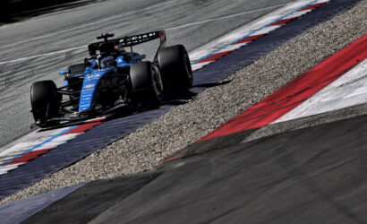Blue Formula 1 race car rounds a corner on a track, riding the red, white, and blue curb with gravel runoff nearby.