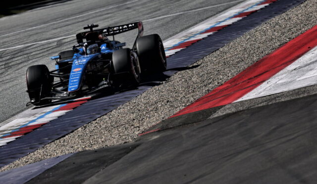 Blue Formula 1 race car rounds a corner on a track, riding the red, white, and blue curb with gravel runoff nearby.