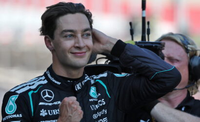 Smiling man in a Mercedes racing suit stands as a cameraman films him in the background.