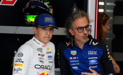 Two men in Oracle Red Bull Racing gear pose in a garage, one in a white suit with cap and the other in a blue suit wearing glasses nearby.