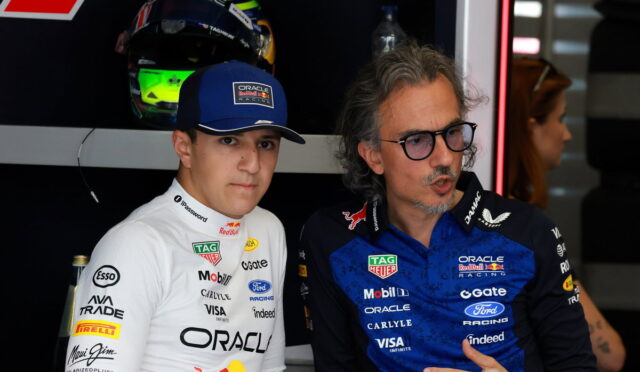 Two men in Oracle Red Bull Racing gear pose in a garage, one in a white suit with cap and the other in a blue suit wearing glasses nearby.