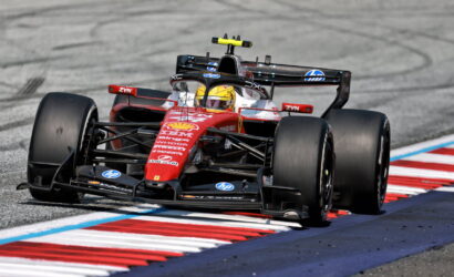 Red Ferrari Formula 1 car navigating a high-speed corner on a race track, driver wearing a yellow helmet with sponsorship logos visible on the car's nose and wings.