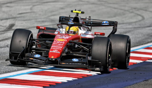 Red Ferrari Formula 1 car navigating a high-speed corner on a race track, driver wearing a yellow helmet with sponsorship logos visible on the car's nose and wings.