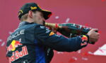 Race car driver in Red Bull uniform sprays champagne from a large bottle in celebration, back to camera, with Oracle logo visible on suit.