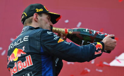 Race car driver in Red Bull uniform sprays champagne from a large bottle in celebration, back to camera, with Oracle logo visible on suit.