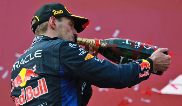 Race car driver in Red Bull uniform sprays champagne from a large bottle in celebration, back to camera, with Oracle logo visible on suit.