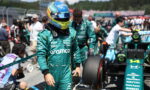 Formula 1 driver in teal Aramco racing suit and blue helmet walks in the pit lane, car and crew in the background.