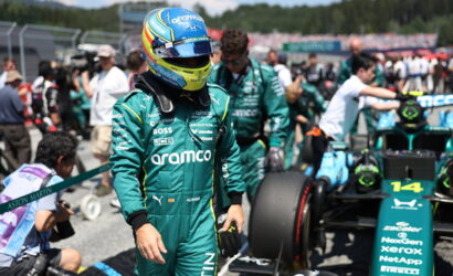 Formula 1 driver in teal Aramco racing suit and blue helmet walks in the pit lane, car and crew in the background.