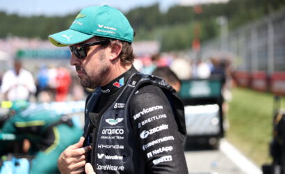 Race car driver wearing a teal cap, sunglasses, and a black sponsor-covered racing suit in the pit lane, adjusting his jacket.