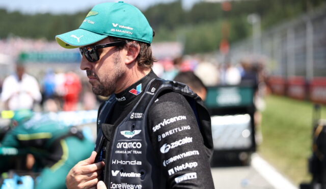 Race car driver wearing a teal cap, sunglasses, and a black sponsor-covered racing suit in the pit lane, adjusting his jacket.