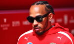Man with braided hair and a beard wearing black sunglasses and a red Ferrari hoodie against a red backdrop, looking downward.