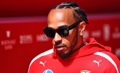 Man with braided hair and a beard wearing black sunglasses and a red Ferrari hoodie against a red backdrop, looking downward.