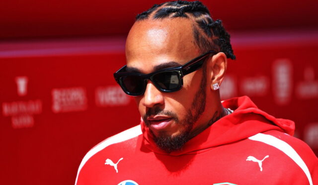 Man with braided hair and a beard wearing black sunglasses and a red Ferrari hoodie against a red backdrop, looking downward.