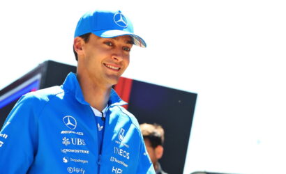 Young man in blue racing team jacket and cap with Mercedes logos, smiling outdoors.