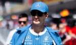 Man wearing a blue Mercedes race team cap and jacket with sponsor logos, sunglasses, in a crowded outdoor event.
