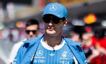 Man wearing a blue Mercedes race team cap and jacket with sponsor logos, sunglasses, in a crowded outdoor event.