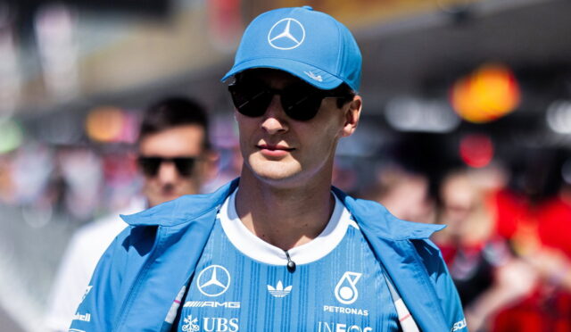 Man wearing a blue Mercedes race team cap and jacket with sponsor logos, sunglasses, in a crowded outdoor event.